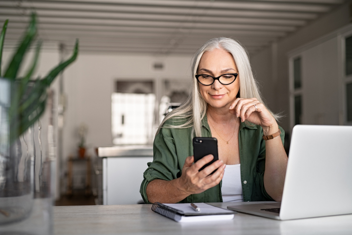 Image of a women sitting at her desk with a notepad, mac book and phone in her hand.