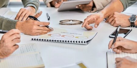 Tips to tackle your debt in 2024 feature view of the hands of five people sitting around a table with a notebook with figures on in the centre of the table
