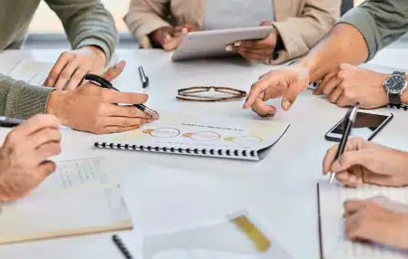 view of the hands of five people sitting around a table with a notebook with figures on in the centre of the table