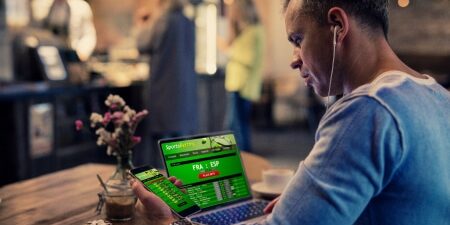 Are-gambling-debts-enforceable_-Feature man sat at laptop with mobile in hand. Both device screens display a gambling app