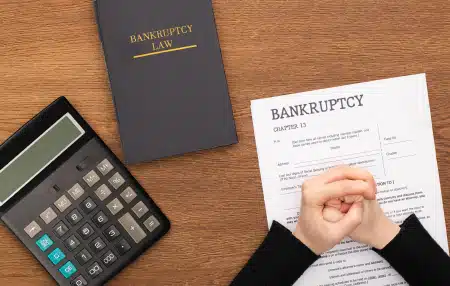 birds eye view of brown wooden table with calculator, black book, and bankruptcy document with persons clasped hands placed on top