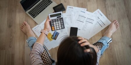 A-list-of-bills-increasing-in-April-feature Woman sat on the wooden floor with lots of paper bills, laptop and calculator.