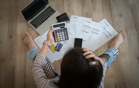 Woman sat on the wooden floor with lots of paper bills, laptop and calculator.