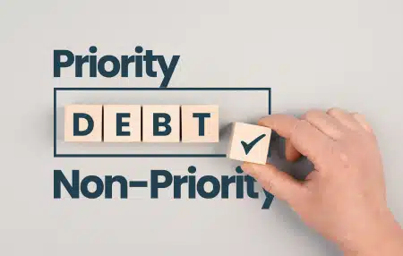 Grey background with the text "Priority Debt and Non-Priority' with some wooden blocks displaying "Debt"