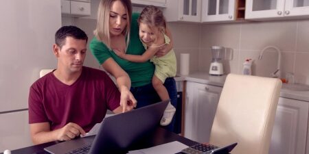 An image of a man and a woman holding a baby in their kitchen, pointing at the laptop looking at their budget.