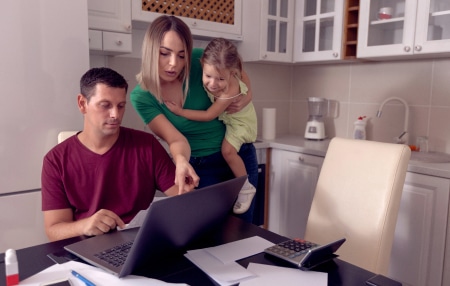 An image of a man and a woman holding a baby in their kitchen, pointing at the laptop looking at their budget.