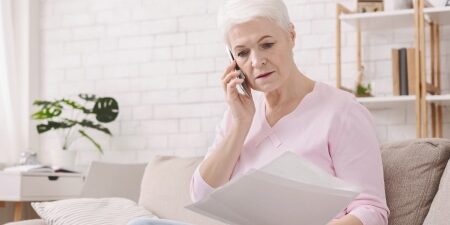 Middle-aged women sitting on her sofa with documents in one hand and speaking on the phone with the other.