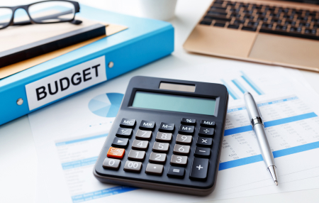 Image of a calculator, pen, laptop, reading glasses and a folder with the label' budget' on.