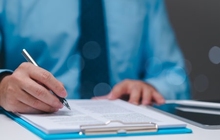 An image of someone wearing a blue shirt with a dark blue tie writing with a pen on a clipboard.