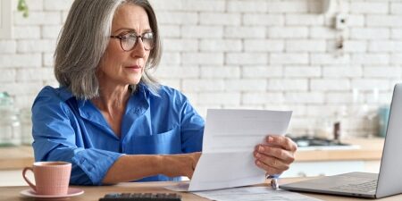 An image of a lady who could be at retirement age, sitting at a desk with a coffee, laptop, and reading forms.