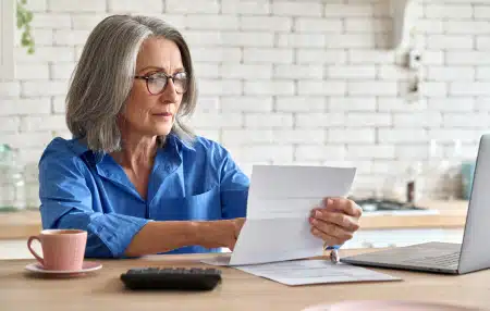 An image of a lady who could be at retirement age, sitting at a desk with a coffee, laptop, and reading forms.