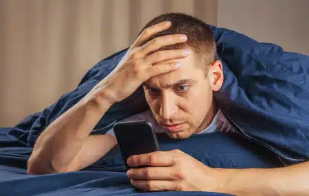 Image of a man lying down in bed on his mobile, looking concerned 