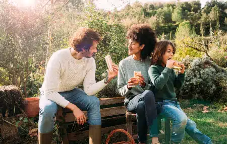 Image of a man and a woman sitting on a bench in the garden with their child, eating sandwiches and smiling