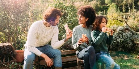 How To Have Fun For Free_ Staying Happy When Dealing with Debt feature Image of a man and a woman sitting on a bench in the garden with their child, eating sandwiches and smiling