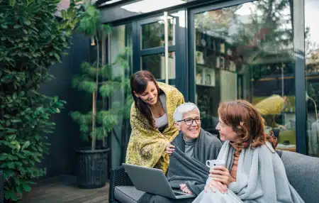 Image of a granddaughter, mother, and grandmother all sitting outside, smiling, with a cup of tea and a laptop. 