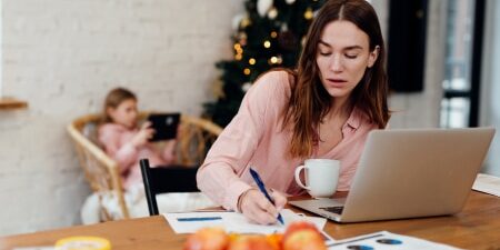 Tips-For-Staying-on-A-budget-feature Image of a mother on her laptop writing her budget with her daughter in the background