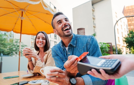 Image of a man and a woman smiling sitting outside of a restaurant paying for their coffee