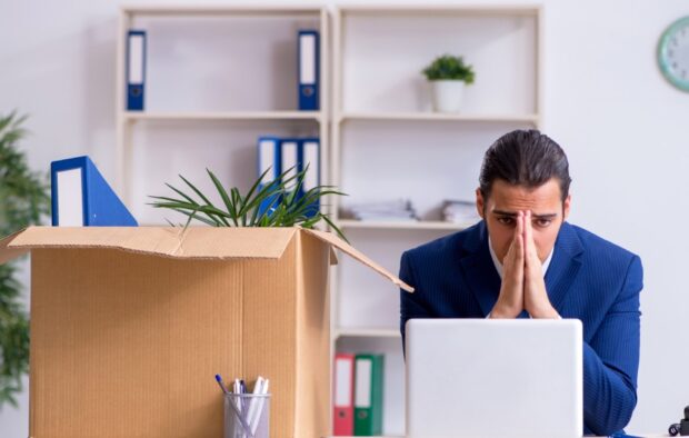 How-To-Cope-With-Job-Loss_-Dealing-With-Debt-After-Redundancy-feature-1 An image of a man sat at his desk with his head in his hands.