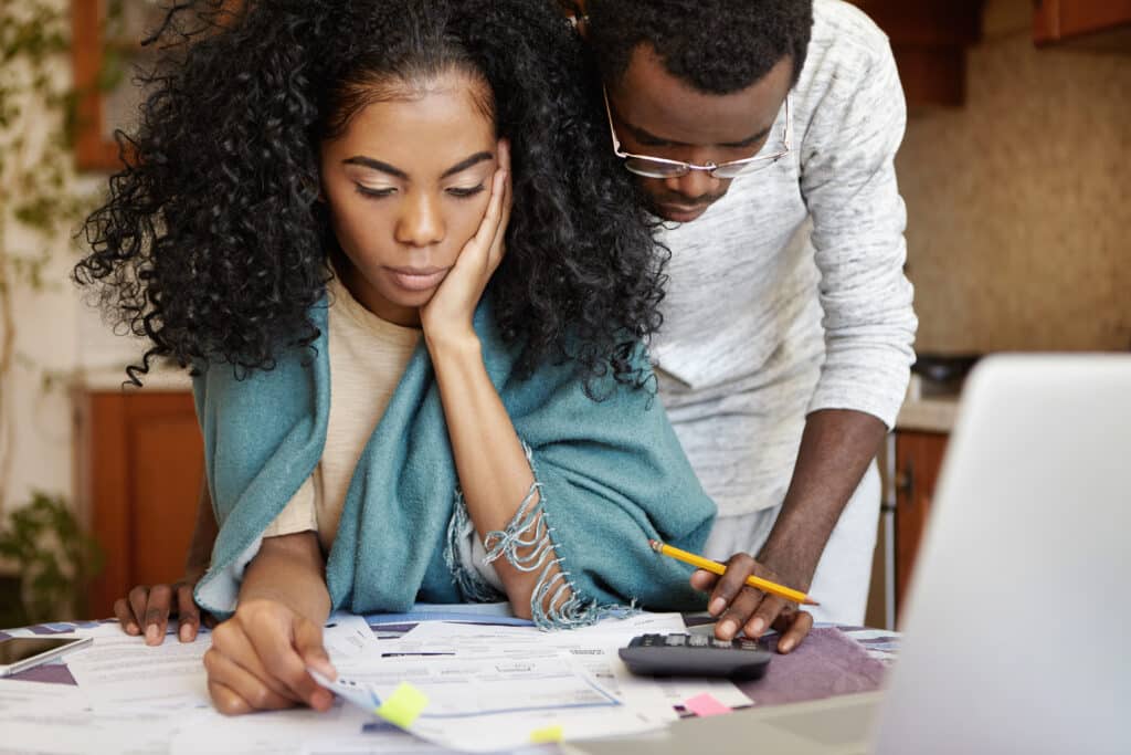 People, family budget, finances and payments concept. African-American man wearing glasses helping his frustrated wife doing paperwork, analysing papers, calculating expenses, using calculator