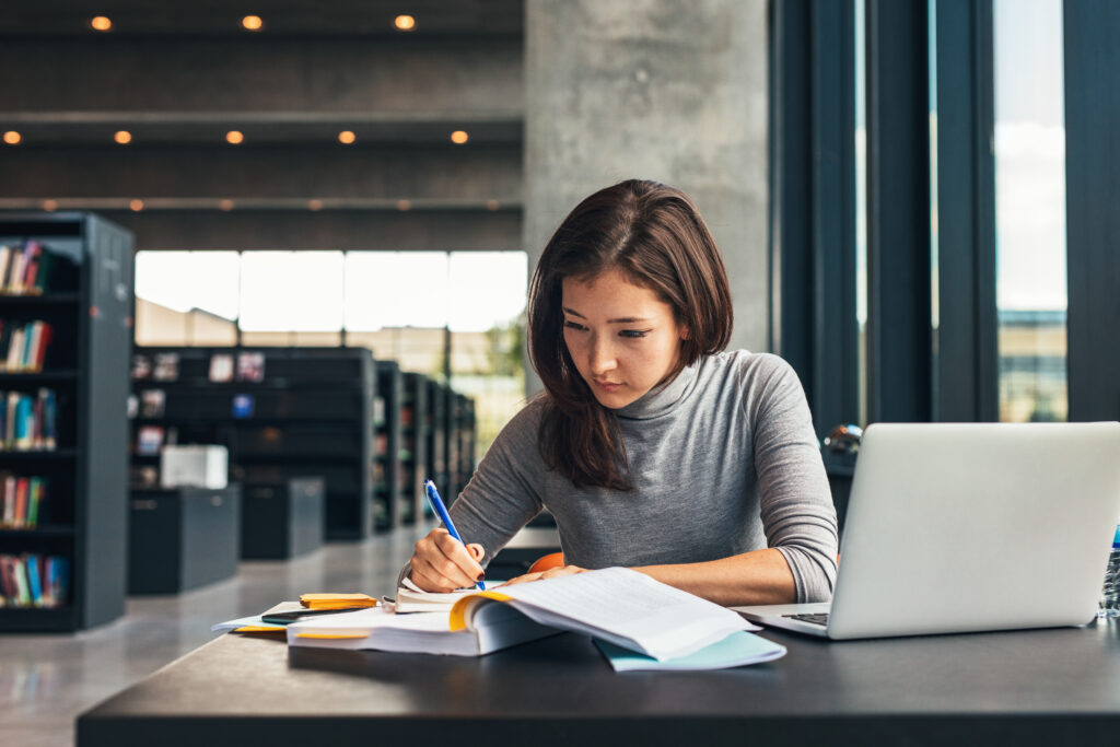 Female student taking notes from a book at library. Young asian woman sitting at table doing assignments in college library.