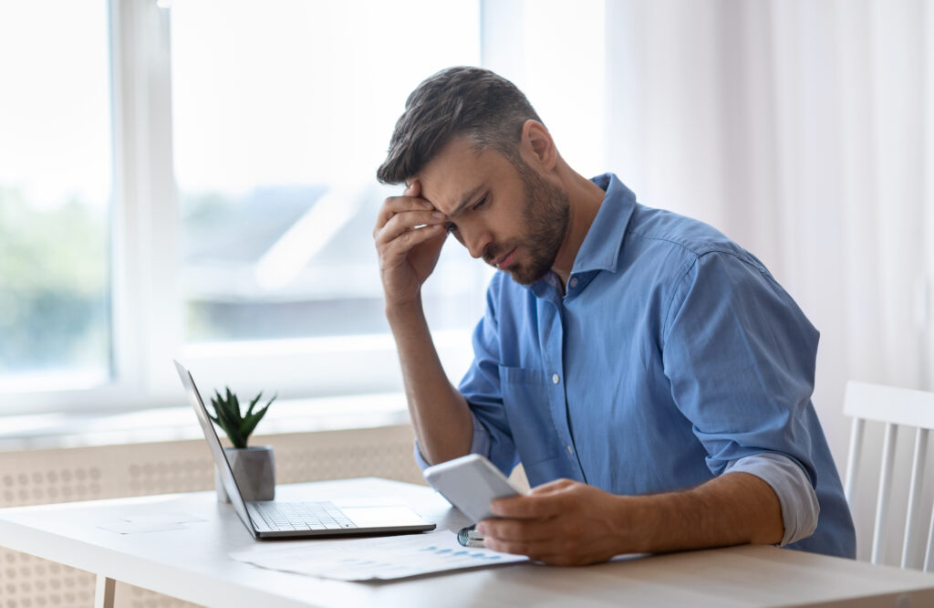 Worried male freelancer looking at smartphone screen while working at home office