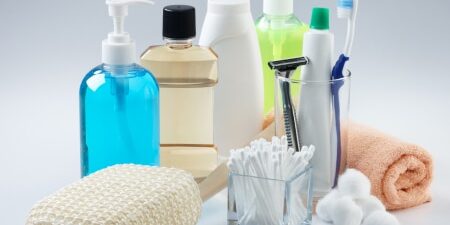 How To Save Money on Toiletries feature An image of toiletries including an orange flannel, blue bottle of liquid soap, mouthwash, shower gel, toothpaste, cotton ear buds, razors and toothpaste.