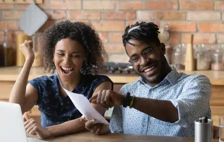 Image of a couple sitting at their table with their laptop, smiling at sheets of paper.