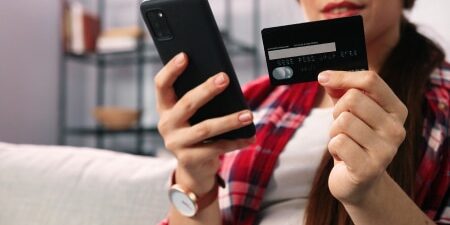 4 Practical Tips for Avoiding Impulse Purchases feature Image of a woman sitting on her sofa with her credit card in one hand and her phone in the other.