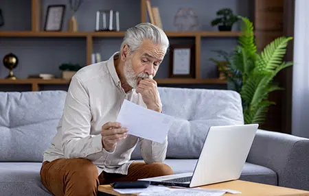 Image of an older man sitting on his sofa reading a letter with his laptop.