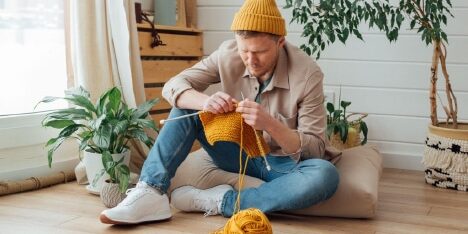 man-knitting A man in a yellow beanie knitting on the floor in a cozy room.