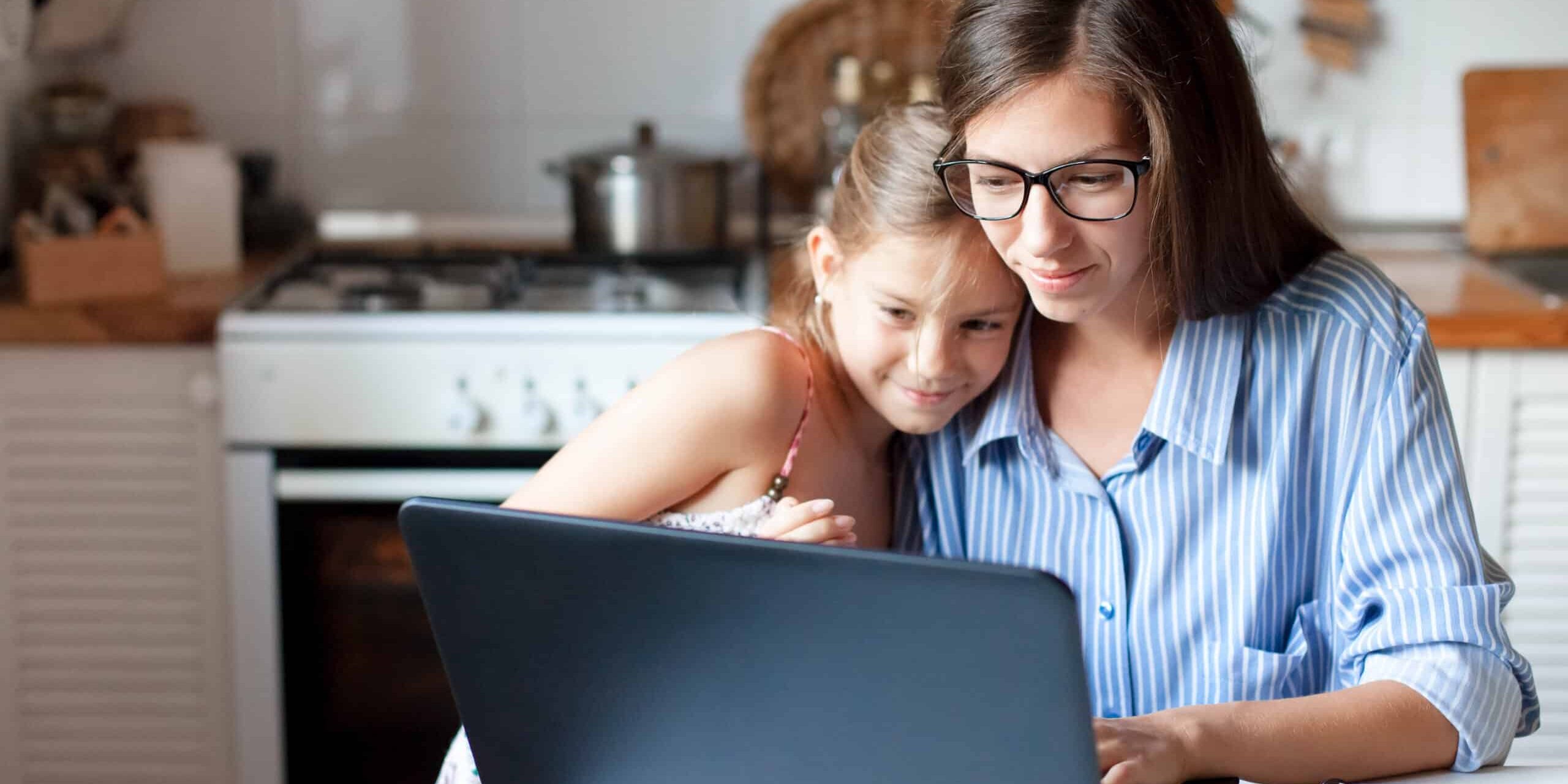 Mother and daughter using laptop and Internet. Freelancer workplace in cozy kitchen. Woman and child A mother using a laptop while her daughter cuddles up to her.