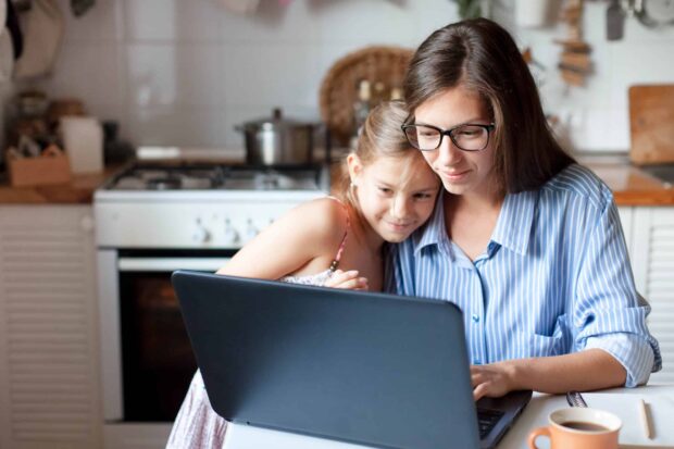 Mother and daughter using laptop and Internet. Freelancer workplace in cozy kitchen. Woman and child A mother using a laptop while her daughter cuddles up to her.