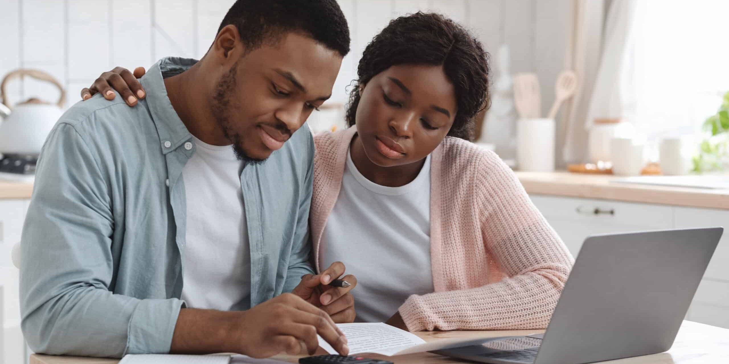 A couple reviews documents together at a kitchen table with a laptop and calculator.