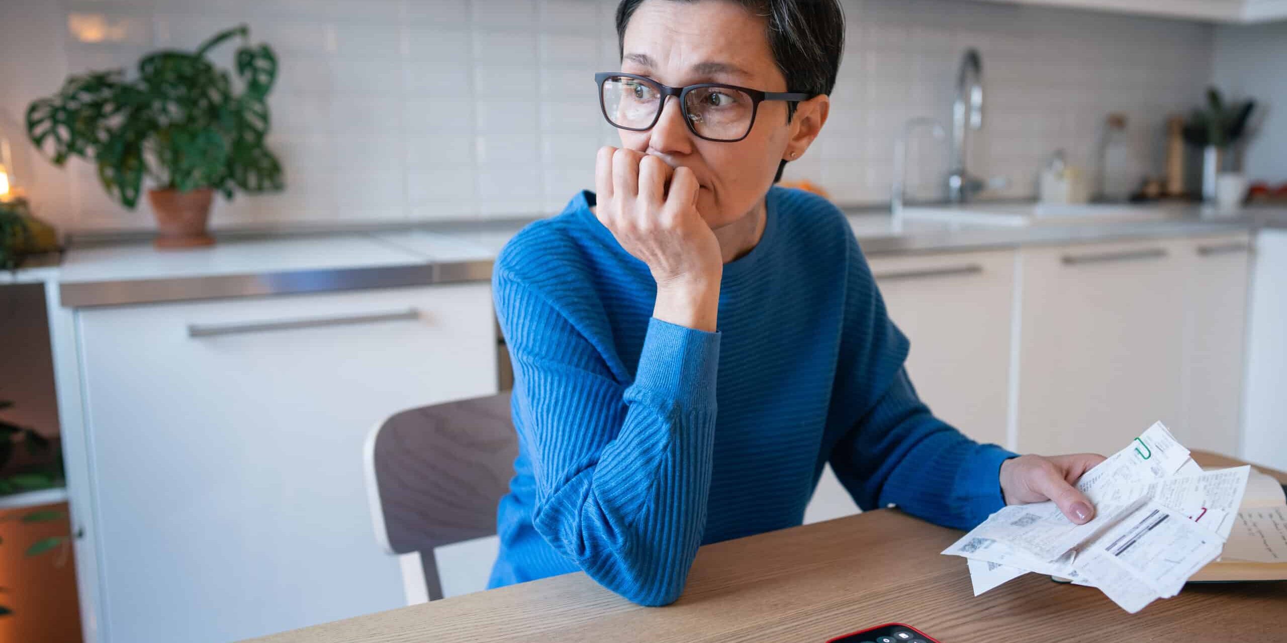 Concerned upset mature woman calculating bills and budget, balancing finances and debts at home. A worried looking woman in a kitchen holding bills, with a calculator app open on her phone.