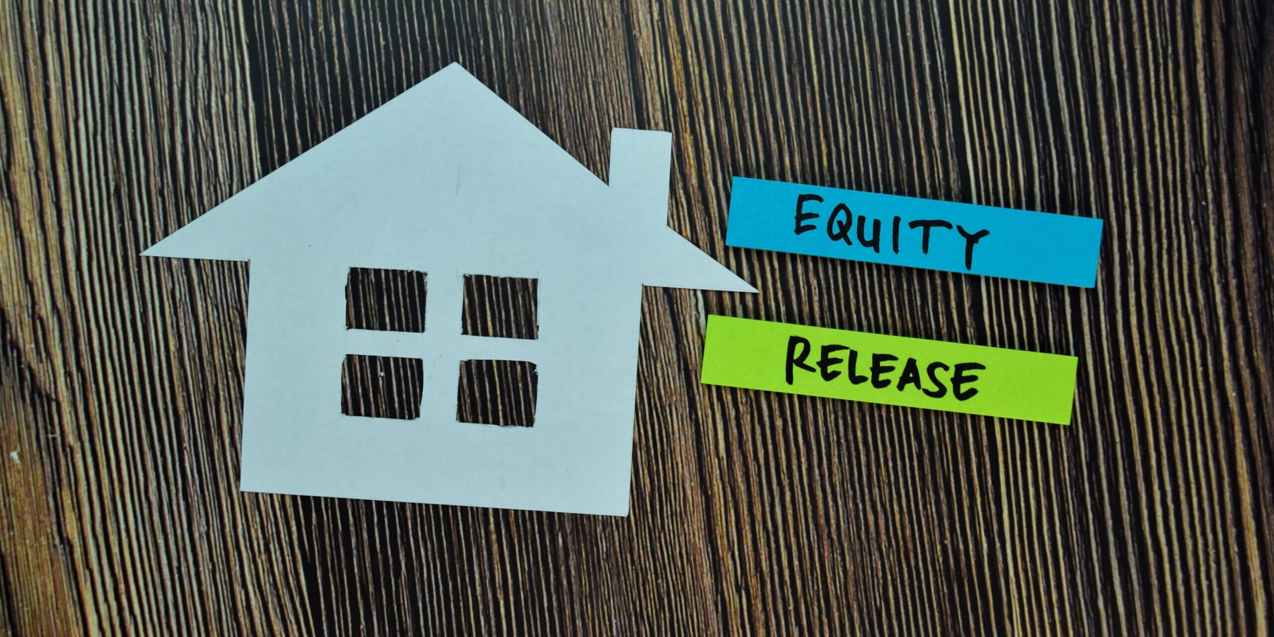Equity Release write on sticky note and house made from paper isolated on Office Desk. Retirement concept A paper house cutout next to labels reading "Equity" and "Release" on a wooden background.
