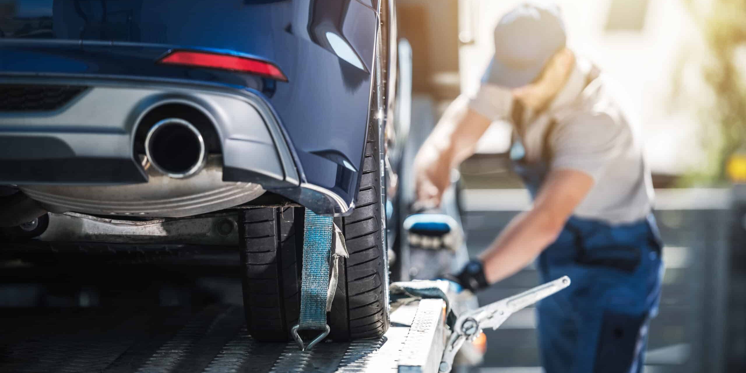 Shipping Brand New Car to Dealership A blue car being secured onto a flatbed tow truck as a worker tightens the strap on the rear tire.