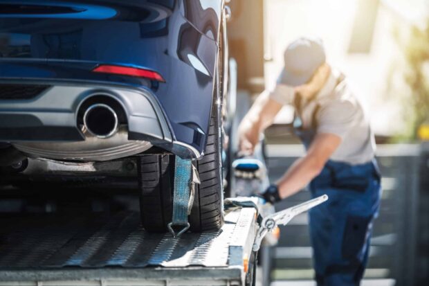 Shipping Brand New Car to Dealership A blue car being secured onto a flatbed tow truck as a worker tightens the strap on the rear tire.