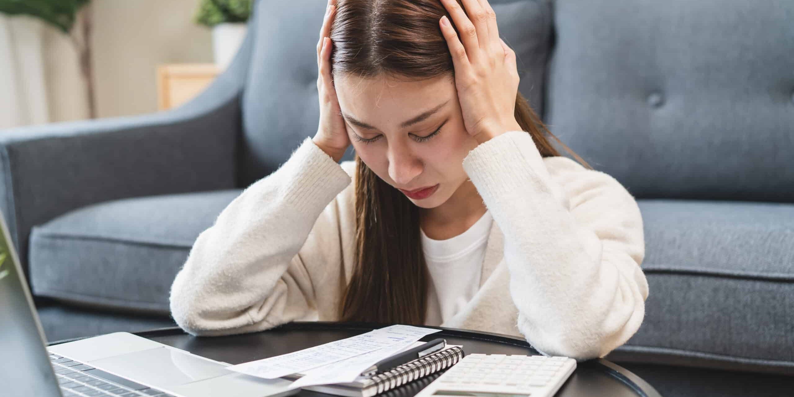 Stressed bankrupt financial crisis concept, Young adult in nervous after calculated loan expense and can not pay to bank. Worried woman looking at bills with a laptop and calculator on the table.