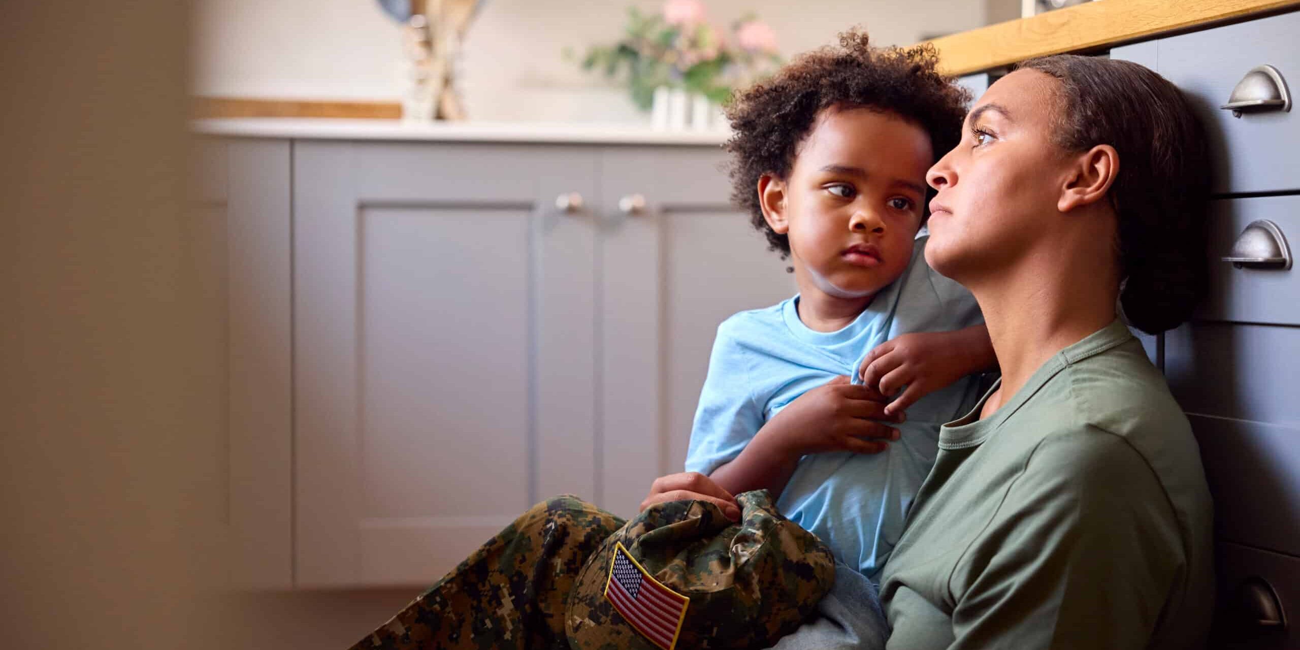 Young Son Comforts Depressed Mother Suffering With PTSD Sitting On Floor On Home Leave