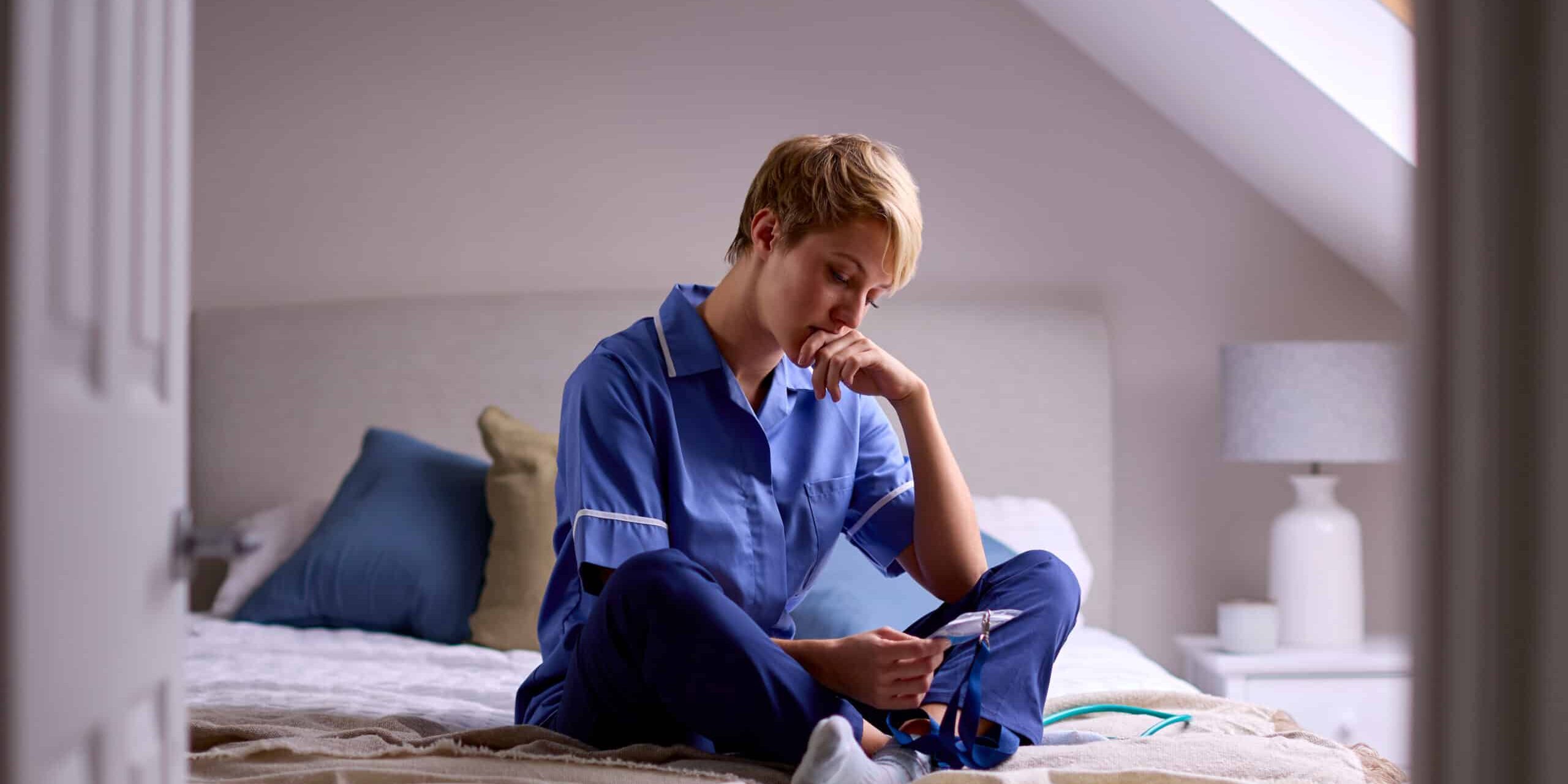 Thoughtful Woman Wearing Nurses Or Doctors Uniform Sitting On Bed At Home Looking At ID Lanyard