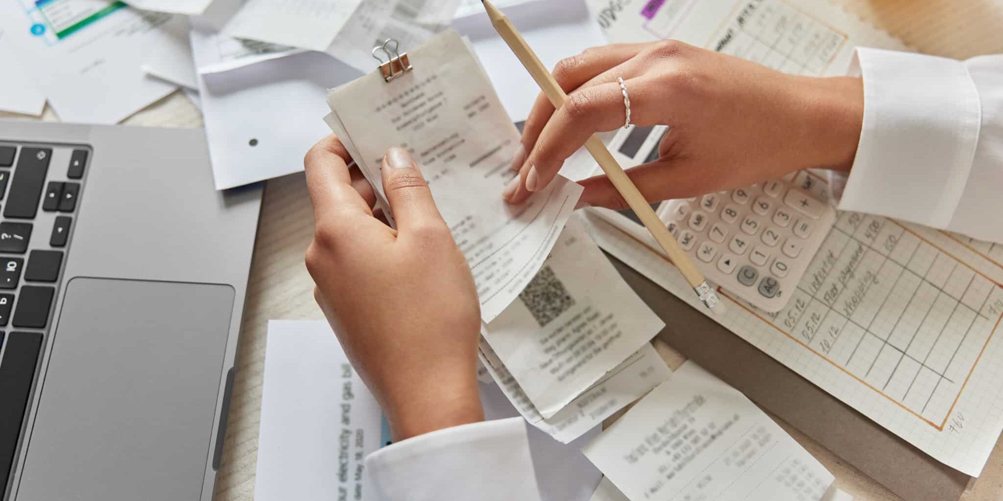 Unrecognizable woman holds receipts pencil uses laptop plans budget manages expenses calculates financial reports checks internet banking service poses at desktop with papers around. View from above