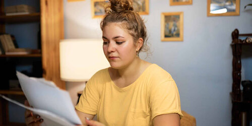 Indoor image of concerned woman in casual yellow t-shirt with hair knob examining paper documents sitting at home in living-room, reading rent agreement or loan statement from bank