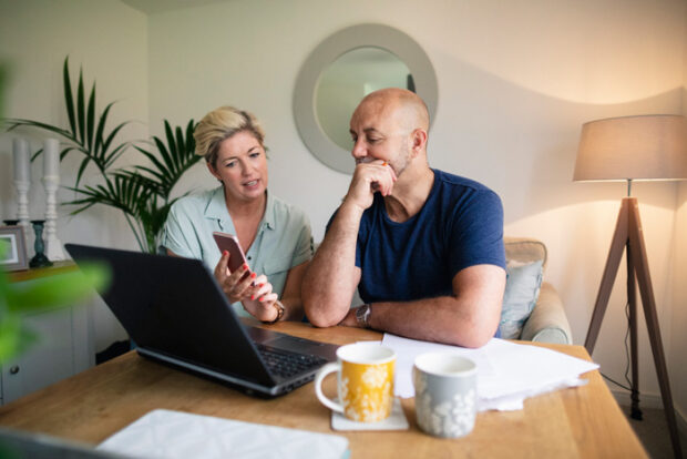 A front-view shot of a happy mature couple enjoying a hot drink while using their laptop, they are wearing casual clothing.